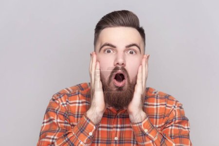 Photo for Portrait of shocked amazed bearded man looking at camera with big eyes and open mouth, keeps palms on cheeks, wearing checkered shirt. Indoor studio shot isolated on gray background. - Royalty Free Image
