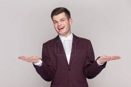 Photo for Portrait of pleased positive optimistic handsome man spreads hands, inviting guests, showing copy space on palms, wearing violet suit and white shirt. Indoor studio shot isolated on grey background. - Royalty Free Image