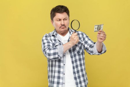 Photo for Portrait of serious concentrated middle aged man in casual checkered shirt looking through loupe to the dollar, checking authenticity of banknote. Indoor studio shot isolated on yellow background. - Royalty Free Image