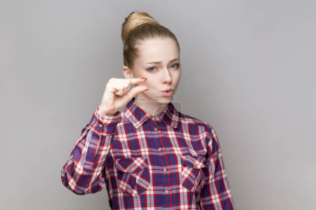 Photo for Portrait of beautiful adorable woman with bun hairstyle standing looking at camera, showing small gesture with fingers, wearing checkered shirt. Indoor studio shot isolated on gray background. - Royalty Free Image