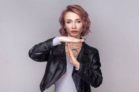 Photo for Portrait of adorable sad woman with short hairstyle standing showing time out gesture, needs more time for her work, wearing black leather jacket. Indoor studio shot isolated on grey background. - Royalty Free Image