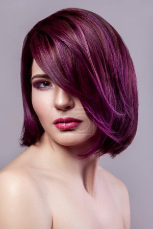 Portrait of beautiful fashion model woman with short purple colored hairstyle and makeup looking at camera with calm expression. Indoor studio shot isolated on gray background.