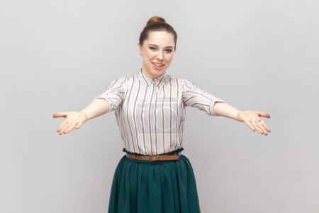 Photo for Please, take it for free. Portrait of pleased friendly optimistic woman wearing striped shirt and green skirt standing with spread hands. Indoor studio shot isolated on gray background. - Royalty Free Image