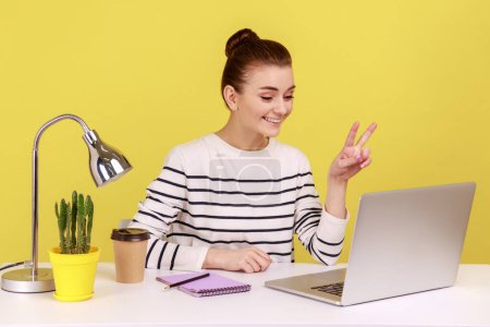 Photo for Positive optimistic woman office manager showing victory gesture and smiling looking into laptop screen, talking video call, online conference. Indoor studio studio shot isolated on yellow background. - Royalty Free Image