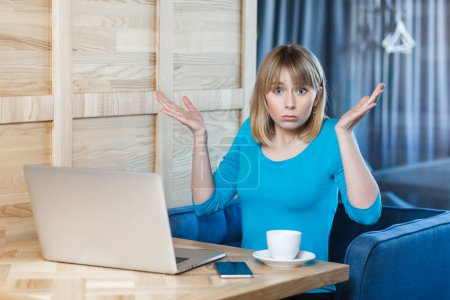 Photo for Confused uncertain young woman with blonde hair in blue shirt sitting at table and working on laptop, shrugging shoulders, looking at camera with puzzled face. Indoor shot, cafe background. - Royalty Free Image