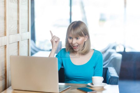 Téléchargez les photos : Smart excité jeune femme positive avec les cheveux blonds en chemise bleue travaillant sur ordinateur portable, levé le doigt, ayant une idée, sait comment résoudre le problème. Intérieur tourné dans un café avec une grande fenêtre sur fond. - en image libre de droit
