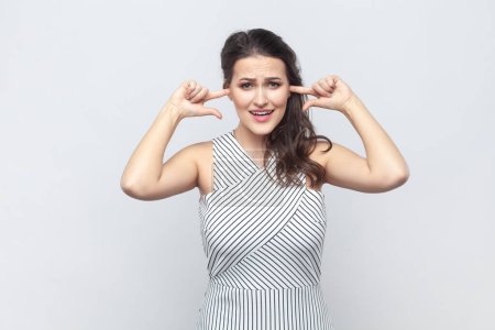 Photo for Portrait of brunette woman covering ears as hears very loud noise or music, clenches teeth, ignores something noisy, wearing striped dress. Indoor studio shot isolated on gray background. - Royalty Free Image