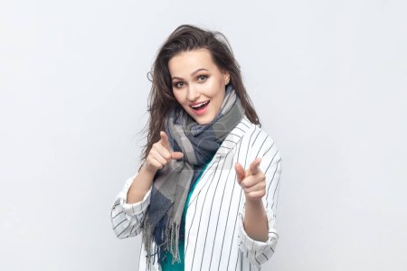 Photo for Portrait of overjoyed happy positive brunette woman pointing at camera with index fingers, choosing you, wearing striped jacket and scarf. Indoor studio shot isolated on gray background. - Royalty Free Image