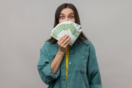 Photo for Amazed happy woman with dark hair hiding face behind fan of euro banknotes, interest-free cash withdrawal, wearing casual style jacket. Indoor studio shot isolated on gray background. - Royalty Free Image