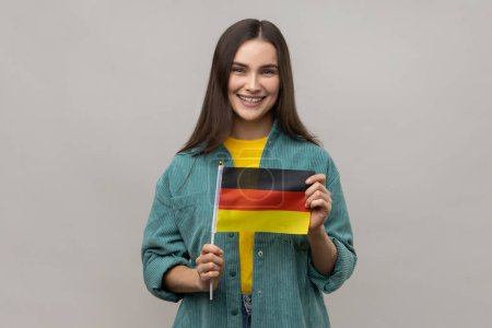 Téléchargez les photos : Portrait de femme séduisante tenant le drapeau de l'Allemagne, célébrant le Jour de l'Allemagne - 3 octobre, exprimant des émotions positives, portant une veste de style décontracté. Studio intérieur tourné isolé sur fond gris. - en image libre de droit