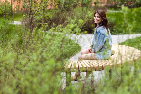 Foto de Retrato de vista lateral de una hermosa mujer con chaqueta de mezclilla sentada en un banco de madera en un parque verde, mirando a la cámara con una sonrisa dentada, disfrutando de su paseo al aire libre. tiro al aire libre. - Imagen libre de derechos