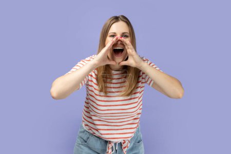 Foto de Mujer rubia adulta joven atractiva enojada que usa una camiseta a rayas de pie y grita con las manos cerca de la boca, expresando emociones agresivas. Estudio interior plano aislado sobre fondo púrpura. - Imagen libre de derechos