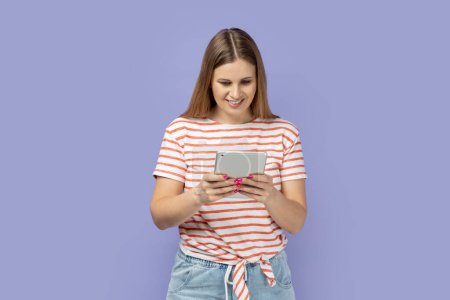 Photo for Portrait of adorable optimistic satisfied woman wearing striped T-shirt holding tablet, reading news or electronic book, looking at display with smile. Indoor studio shot isolated on purple background - Royalty Free Image