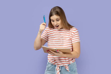 Foto de Retrato de mujer rubia optimista emocionada asombrada que lleva una camiseta rayada escribiendo en un cuaderno de papel, pluma levantada, que tiene una idea excelente. Estudio interior plano aislado sobre fondo púrpura. - Imagen libre de derechos