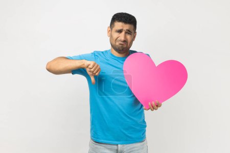 Photo for Portrait of sad upset romantic unshaven man wearing blue T- shirt standing holding big pink heart and showing thumb down, dislike gesture. Indoor studio shot isolated on gray background. - Royalty Free Image