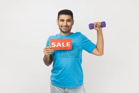 Photo for Portrait of smiling delighted man wearing blue T- shirt standing holding dumbbell and card with sale inscription, discounts for fitness season ticket. Indoor studio shot isolated on gray background - Royalty Free Image