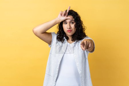 Photo for Portrait of grumpy disrespectful woman with dark wavy hair showing loser gesture and pointing finger at camera, blaming and mocking. Indoor studio shot isolated on yellow background. - Royalty Free Image
