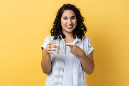 Photo for Portrait of smiling happy positive beautiful woman with dark wavy hair standing with glass of water, pointing at beverage with finger. Indoor studio shot isolated on yellow background. - Royalty Free Image