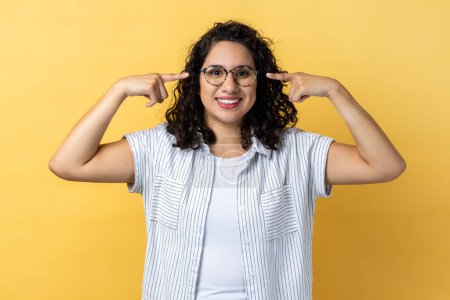 Photo for Portrait of smiling friendly positive woman with dark wavy hair in eyeglasses, standing pointing at her new spectacles, looking at camera. Indoor studio shot isolated on yellow background. - Royalty Free Image