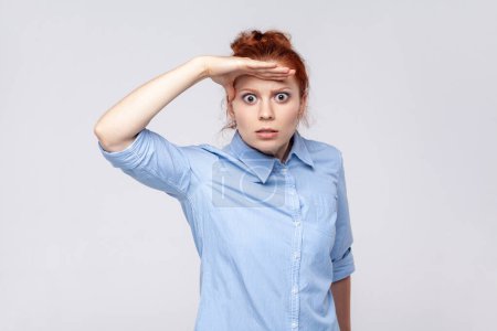 Photo for Portrait of scared ginger woman wearing blue shirt keeping palm over head and looking attentively far away, peering at long distance. Indoor studio shot isolated on gray background. - Royalty Free Image