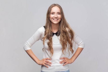 Photo for Portrait of happy joyful cheerful beautiful woman with long blond hair standing with hands on hips, looking at camera, expressing happiness. Indoor studio shot isolated on gray background. - Royalty Free Image