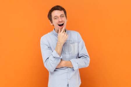 Photo for Portrait of excited amazed young adult man standing with hand on chin, having good idea, looking at camera, wearing light blue shirt. Indoor studio shot isolated on orange background. - Royalty Free Image