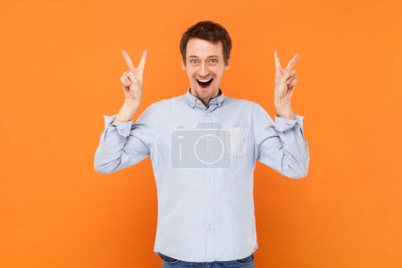 Téléchargez les photos : Portrait de jeune homme optimiste satisfait debout montrant le signe v, regardant la caméra, célébrant la victoire, portant une chemise bleu clair. Studio intérieur tourné isolé sur fond orange. - en image libre de droit