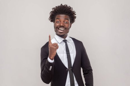 Photo for Clever man with Afro hairstyle get new idea, raises fore finger, ready to start work on project, confident in success, wearing white shirt and tuxedo. Indoor studio shot isolated on gray background. - Royalty Free Image