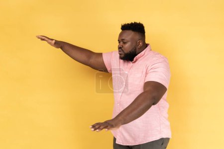 Photo for Side view of lonely blind disabled man wearing pink shirt standing with closed eyes and outstretched hands searching way, walking with doubts. Indoor studio shot isolated on yellow background. - Royalty Free Image