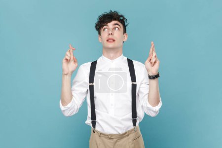 Photo for Portrait of hopeful man wearing white shirt and suspender standing with crossed fingers, waiting for miracle, make a wish. Indoor studio shot isolated on blue background. - Royalty Free Image