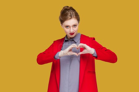 Photo for Portrait of romantic flirting woman with red lips standing showing heart shape with hands, looking at camera, expressing devotion, wearing red jacket. Indoor studio shot isolated on yellow background. - Royalty Free Image