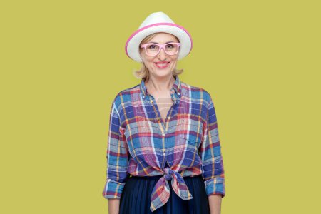 Photo for Portrait of smiling joyful senior woman wearing checkered shirt, hat and eyeglasses, looking at camera with toothy smile, expressing positive emotions. Indoor studio shot isolated on yellow background - Royalty Free Image