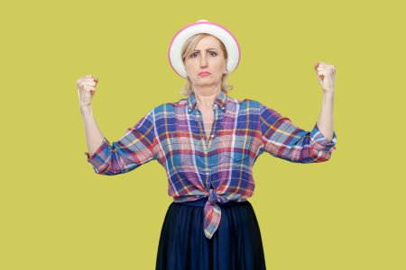 Photo for Portrait of mature woman wearing checkered shirt and hat raises hands and shows muscles, feels proud about her achievements in gym, smiles broadly. Indoor studio shot isolated on yellow background. - Royalty Free Image