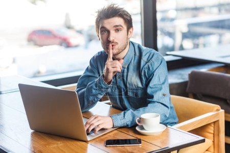 Photo for Portrait of handsome bearded young man freelancer in blue jeans shirt working on laptop, showing shh gesture, looking at camera, asking not to make noise. Indoor shot near big window, cafe background. - Royalty Free Image