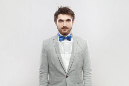 Photo for Portrait of calm positive bearded man standing looking at camera, having relaxed facial expression, wearing grey suit and blue bow tie. Indoor studio shot isolated on gray background. - Royalty Free Image