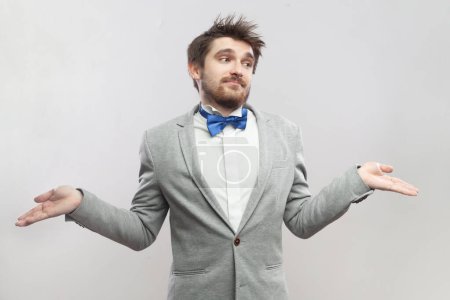 Photo for Portrait of uncertain handsome bearded man spreads hands, shrugging shoulders, doesn't know how to find solution, wearing grey suit and blue bow tie. Indoor studio shot isolated on gray background. - Royalty Free Image