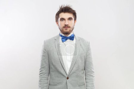 Photo for Portrait of serious attentive concentrated bearded man standing looking at camera with strict bossy expression, wearing grey suit and blue bow tie. Indoor studio shot isolated on gray background. - Royalty Free Image