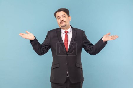 Photo for Portrait of confused uncertain man with mustache with spread hands, looking at camera, shrugging shoulders, wearing black suit with red tie. Indoor studio shot isolated on light blue background. - Royalty Free Image