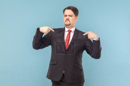 Portrait of bossy confident man pointing at himself with both index fingers, looking at camera with frowning face, wearing black suit with red tie. Indoor studio shot isolated on light blue background