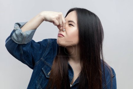 Portrait of disgusted brunette woman in blue denim jacket standing plugs nose as smells something stink and unpleasant, feels aversion. Indoor studio shot isolated on gray background.