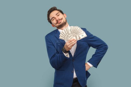 Photo for Portrait of rich proud satisfied man with mustache standing showing fan of dollars banknotes, keeps hand on hip, wearing official style suit. Indoor studio shot isolated on light blue background. - Royalty Free Image
