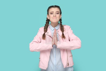 Téléchargez les photos : Portrait d'adolescente pleine d'espoir avec des tresses portant une veste rose debout garde la main dans le geste de prière, suppliant, demandant de pardonner. Studio intérieur tourné isolé sur fond vert. - en image libre de droit