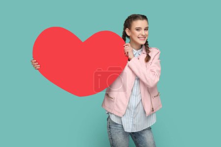 Photo for Portrait of extremely joyful cheerful teenager girl with braids wearing pink jacket, showing big red heart, smiling to camera. Indoor studio shot isolated on green background. - Royalty Free Image