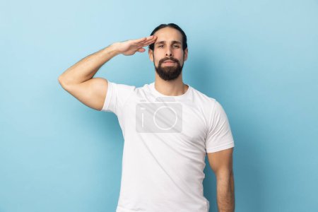 Photo for Portrait of serious responsible man bodybuilder with beard giving salute listening to order with serious attentive expression, following discipline. Indoor studio shot isolated on blue background. - Royalty Free Image