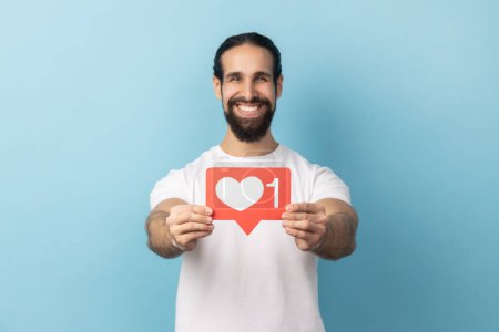 Téléchargez les photos : Portrait d'homme barbu portant un T-shirt blanc tenant le cœur comme une icône des médias sociaux et regardant la caméra avec un sourire de dents. Studio intérieur tourné isolé sur fond bleu. - en image libre de droit