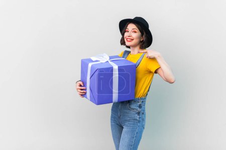 Photo for Portrait of smiling pretty hipster woman in blue denim overalls, yellow T-shirt and black hat holding pointing at big present box with gift for birthday. Indoor studio shot isolated on gray background - Royalty Free Image