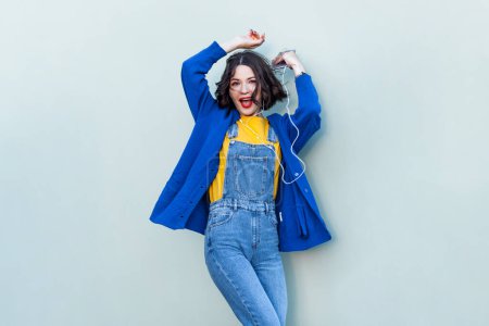 Photo for Portrait of happy cheerful hipster woman in denim overalls, yellow T-shirt and blue jacket, listening to music, dancing with raised hands. Indoor studio shot isolated on light green background. - Royalty Free Image