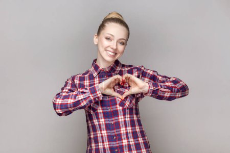 Photo for Portrait of pleased friendly woman with bun hairstyle looking at camera with charming smile, showing heart shape gesture, wearing checkered shirt. Indoor studio shot isolated on gray background. - Royalty Free Image