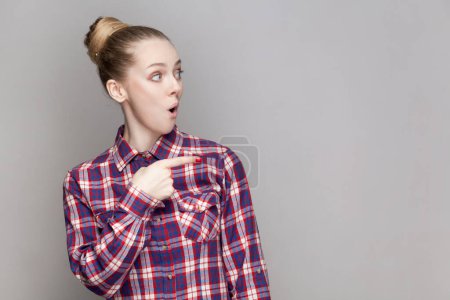 Photo for Portrait of amazed surprised woman with bun hairstyle looking at pointing away with shocked expression, advertisement area, wearing checkered shirt. Indoor studio shot isolated on gray background. - Royalty Free Image