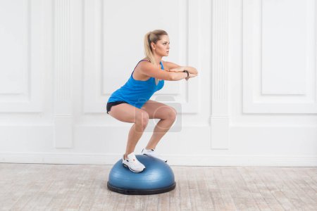 Photo for Side view portrait of sporty attractive woman wearing black shorts and blue top working in gym doing exercise in bosu balance trainer, squats on fitness ball, holding balance. Indoor studio shot. - Royalty Free Image